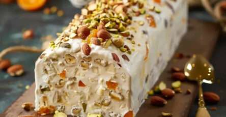 Tranche de nougat glacé maison aux fruits secs, parsemée d’amandes caramélisées, pistaches et abricots, servie avec un coulis de fruits rouges sur une assiette de fête.