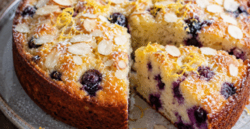 Gâteau myrtilles citron moelleux avec amandes effilées, tranches de citron et myrtilles fraîches sur une table rustique.